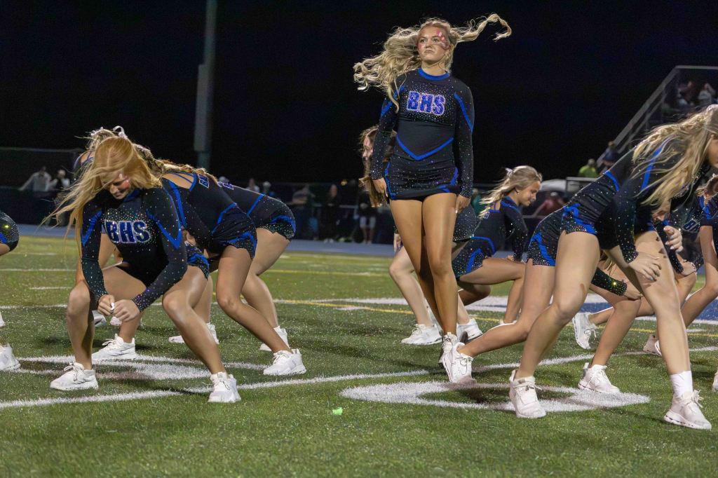 A group of cheerleaders performing a synchronized routine on a football field at night, wearing black and blue uniforms with sparkles.