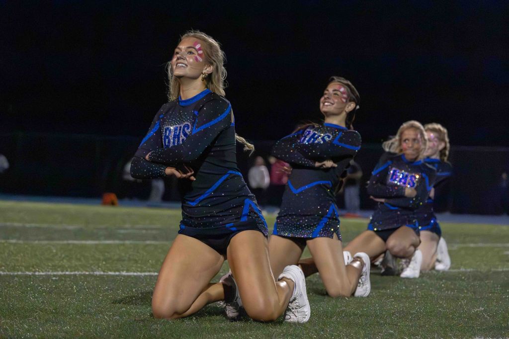 A group of cheerleaders performing a dance routine on a football field at night, wearing black and blue cheer uniforms with glitter accents and face paint.