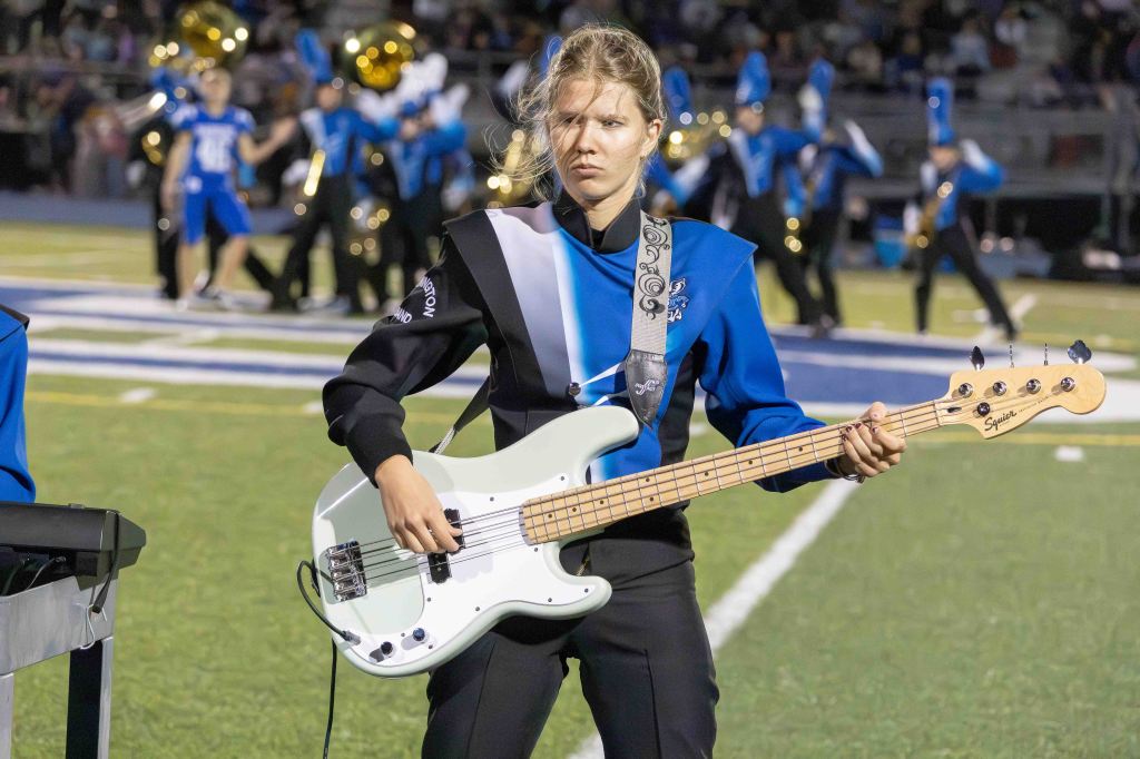 A female musician wearing a blue and black marching band uniform plays an electric bass guitar on a football field, with additional band members performing in the background.