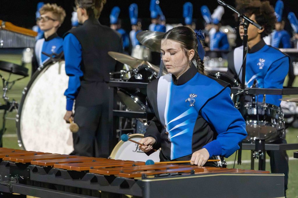 A young female percussionist playing a xylophone, wearing a blue and black marching band uniform, while fellow band members in the background are performing.