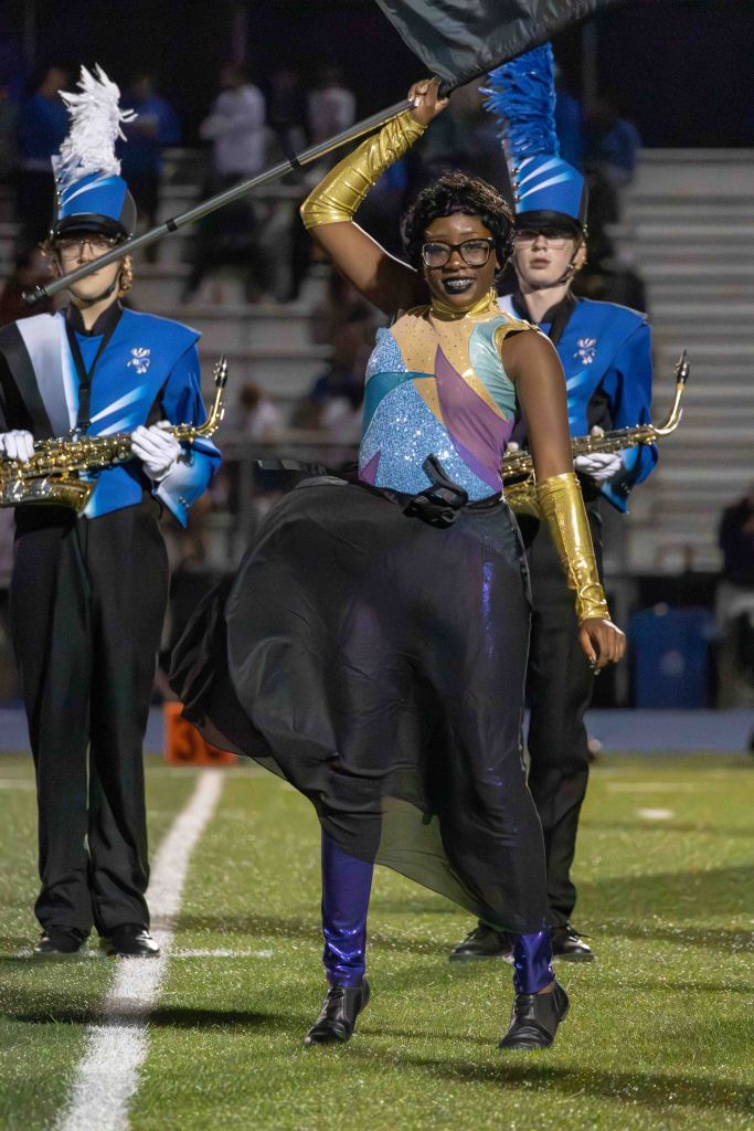 A performer in colorful attire dances while holding a flag during a marching band performance on a football field.