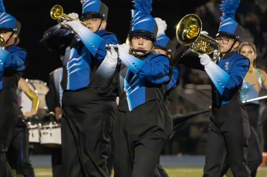 A marching band performing on a field at night, with members wearing blue and black uniforms. A musician plays a flute while others hold various instruments, contributing to the band ensemble.