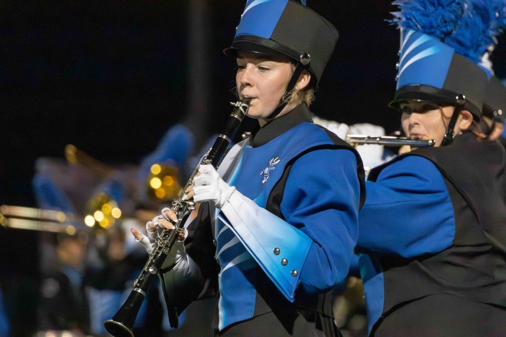A musician in a blue and black marching band uniform plays the clarinet, with a focused expression. In the background, other band members are visible, also in uniform.