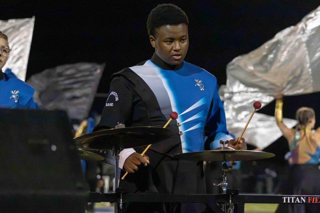 A drummer in a blue and black uniform playing a drum set during a marching band performance at night. A color guard member can be seen in the background.
