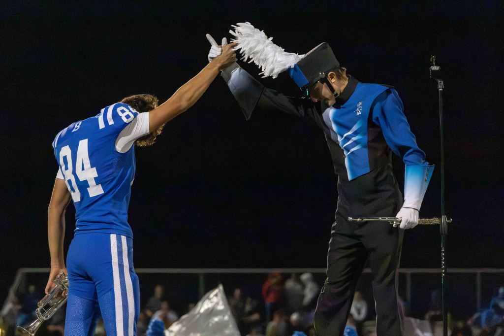 A football player in a blue jersey, number 84, kneels while holding a trumpet as a band conductor in a blue and black uniform raises a white plumed hat above him, celebrating a moment during a performance.