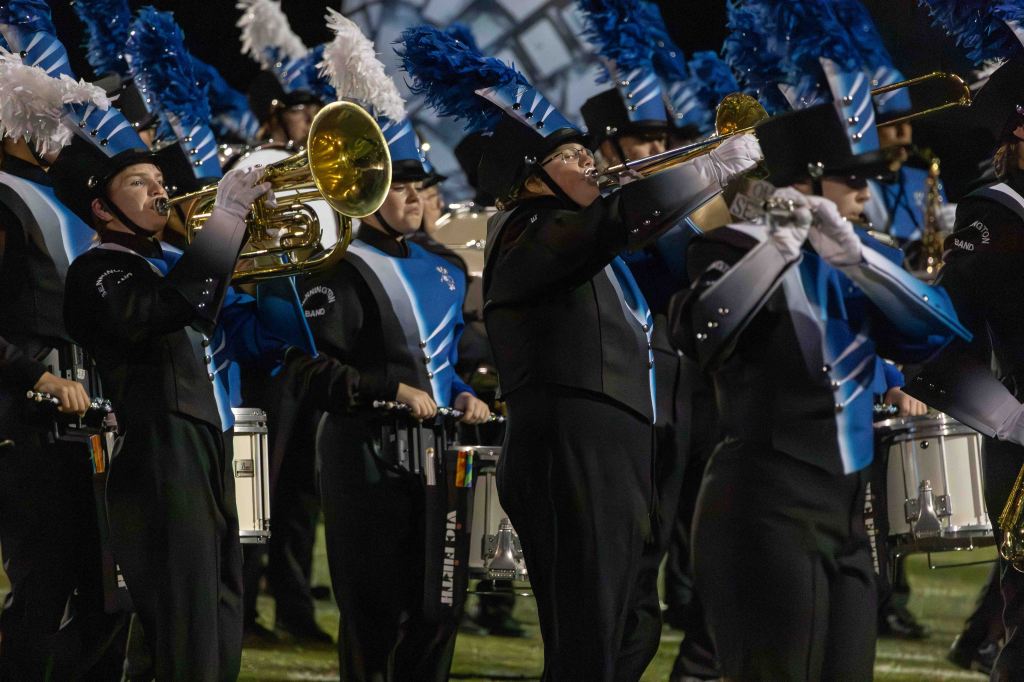 A marching band performing during a night event, featuring musicians playing brass instruments and drums, dressed in black and blue uniforms with feathered hats.