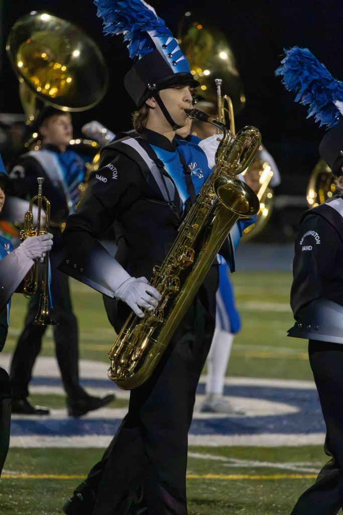 Marching band member playing a saxophone, wearing a black uniform with blue accents and a tall, feathered hat.