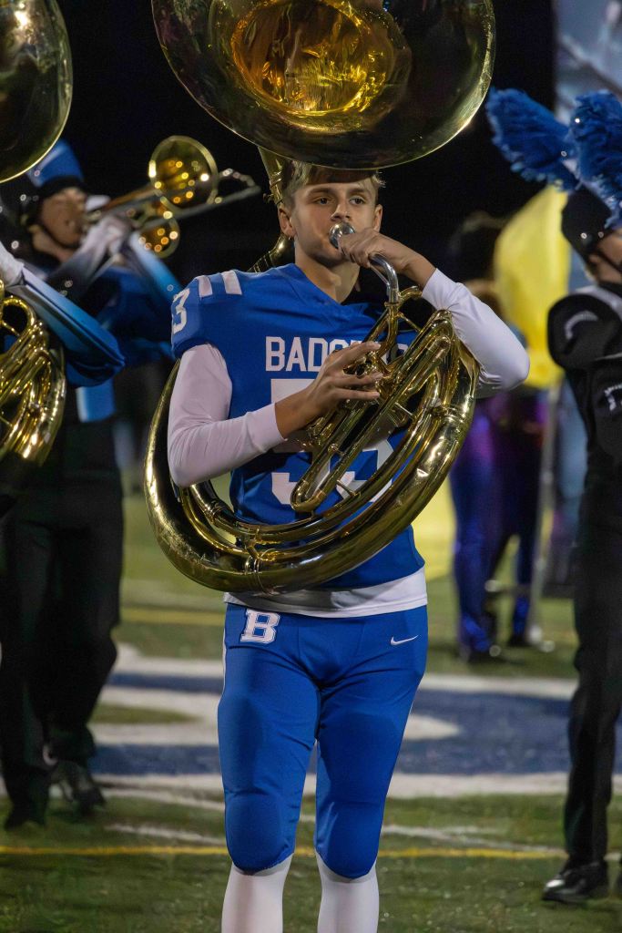 A young musician in a blue football jersey playing a tuba on a sports field, surrounded by fellow band members.
