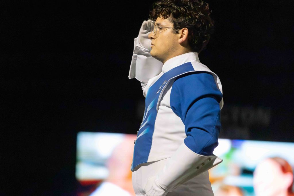 A marching band member in a blue and white uniform saluting on stage.