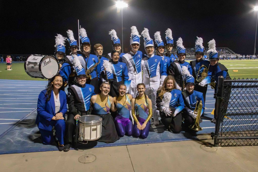 A marching band posed for a group photo on a sports field at night, with members in blue and white uniforms, some holding instruments. The band includes musicians and color guard members, all smiling.