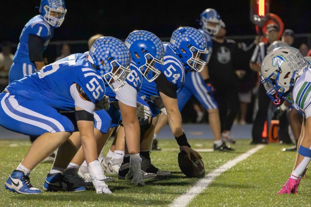 A football team in blue uniforms prepares to snap the ball, lined up on the field, facing an opposing player in a white and green uniform.