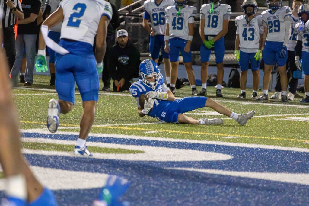 A high school football player in blue and white uniform successfully catches the ball while diving on the field, with teammates and spectators in the background.