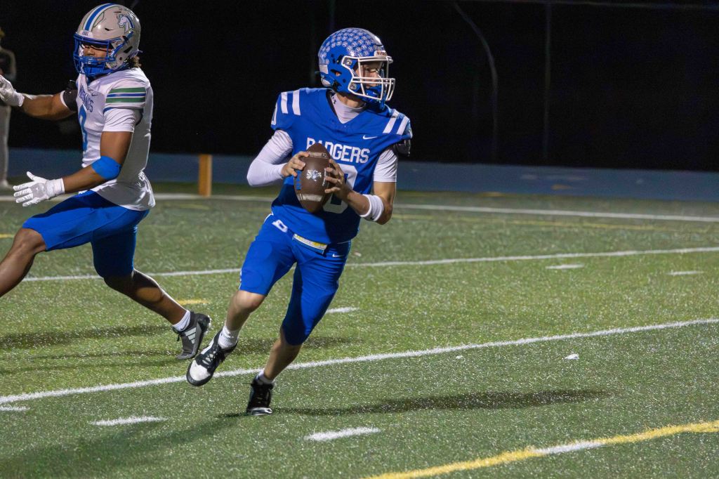 A football player in a blue jersey, with 'Badgers' written on it, spins away from a defender while preparing to throw the ball during a night game.