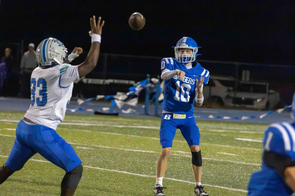 A football player in a blue jersey with the number 10 is preparing to throw a football while another player wearing a white jersey with the number 33 is attempting to block the pass. The scene is set on a football field at night.
