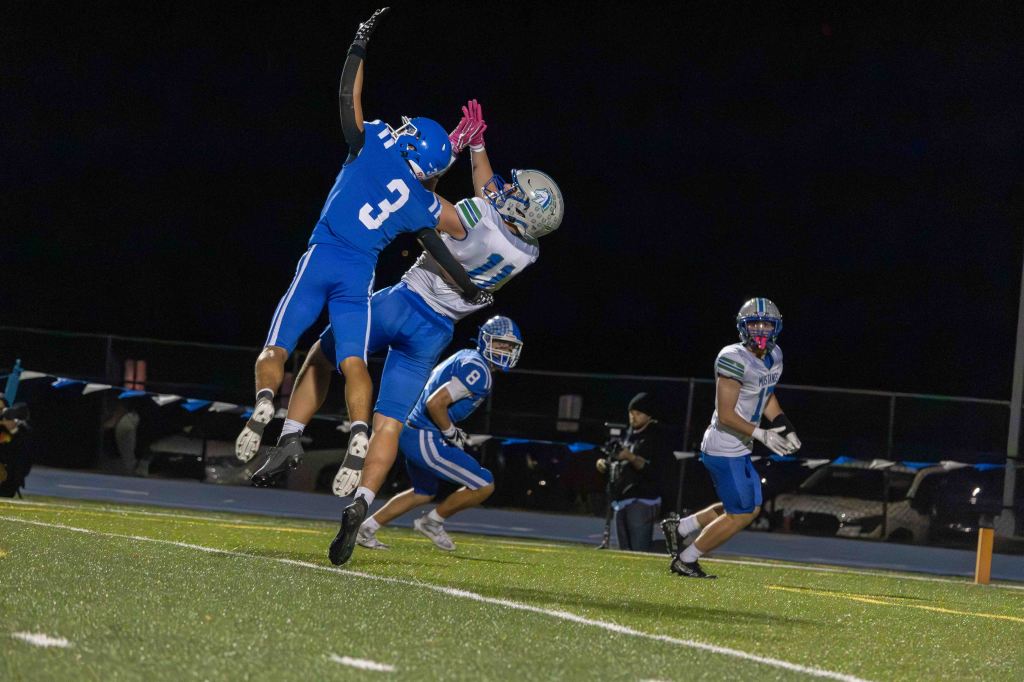 A football player in a blue jersey jumps to intercept a pass while another player in a white jersey attempts a catch during a night game. Other players are in the background.