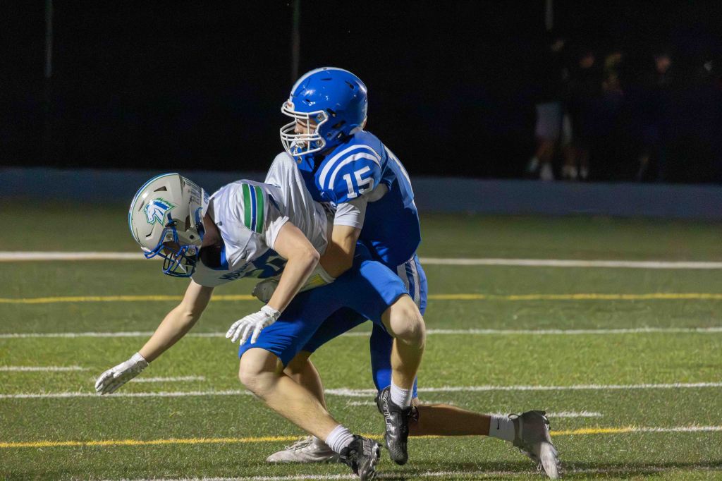A football player in blue tackles an opposing player in a white uniform on a green field during a night game.