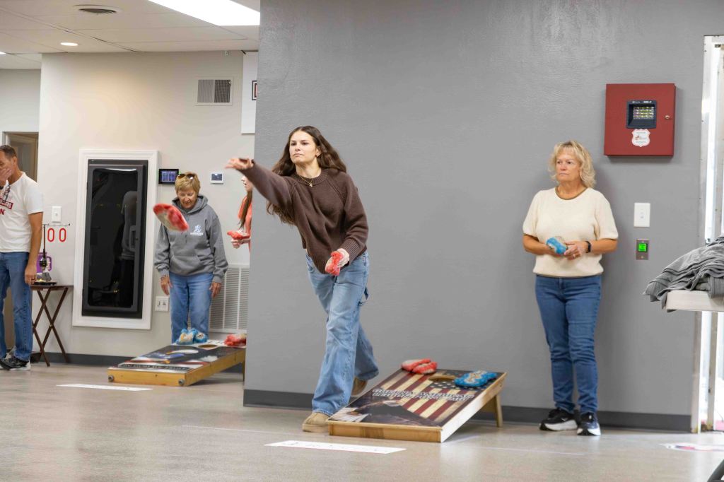 A group of people participating in a cornhole game indoors, with one player throwing a bean bag while others stand nearby waiting for their turn.
