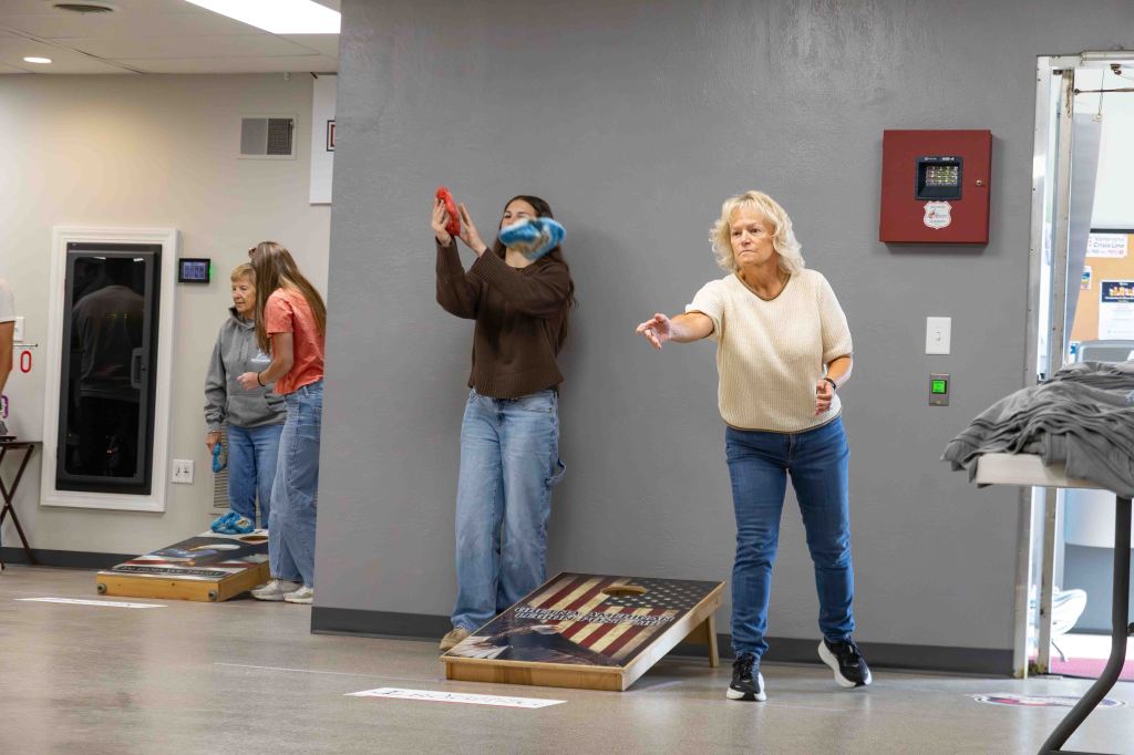 Two women and one girl playing cornhole indoors, with one woman in action throwing a bean bag towards the board.