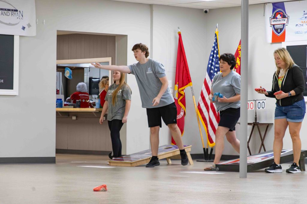 Participants playing a bean bag toss game in a recreation area, with flags and a scoreboard in the background.