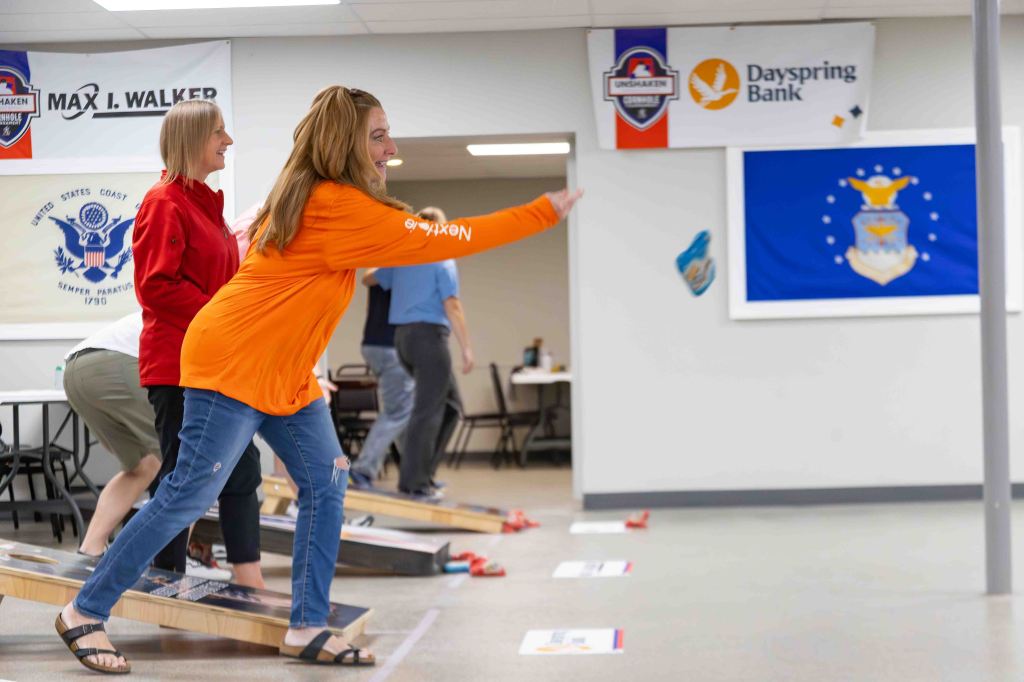 A woman wearing an orange shirt participates in a bean bag toss game, while others play in the background. Various banners are displayed on the walls.