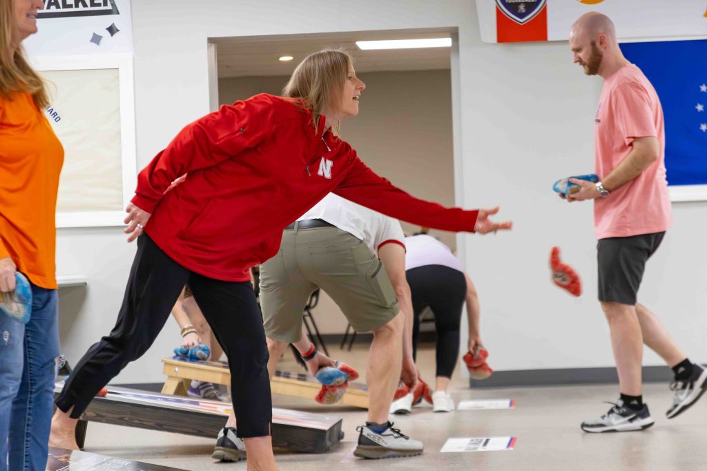 A group of people participating in a game, with one person in a red sweatshirt throwing a bag towards a target. Others are in the background, engaged in the same activity.