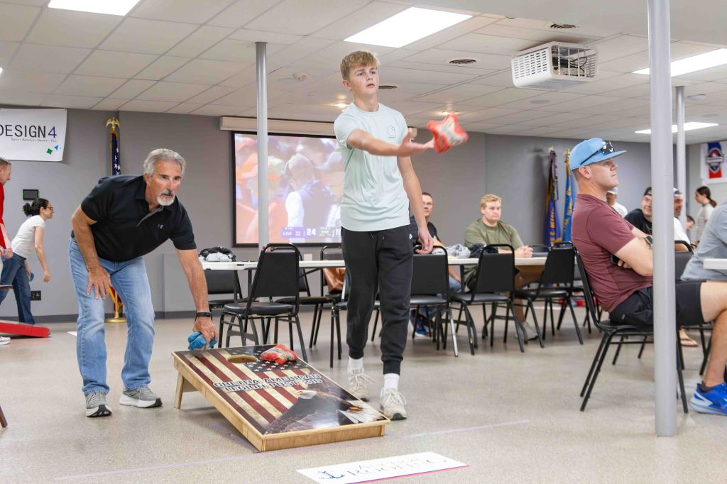 A young male competitor throwing a red cornhole bag towards a wooden board with an American flag design, while an older man prepares to throw a blue bag next to him. In the background, a group of people is seated at tables, with a sports game playing on a screen.