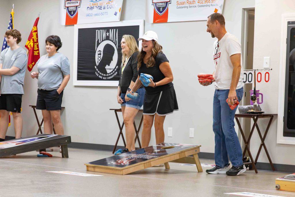 Participants at a cornhole tournament, smiling and preparing to throw bags, with a scoreboard in the background.