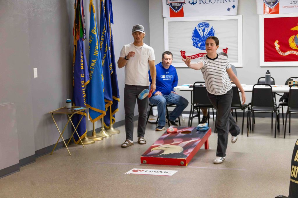 A young girl throws a red cornhole bag while a man watches closely. Another man sits on a chair, observing the game. Flags hang on the wall behind them, and there is a cornhole board on the floor.