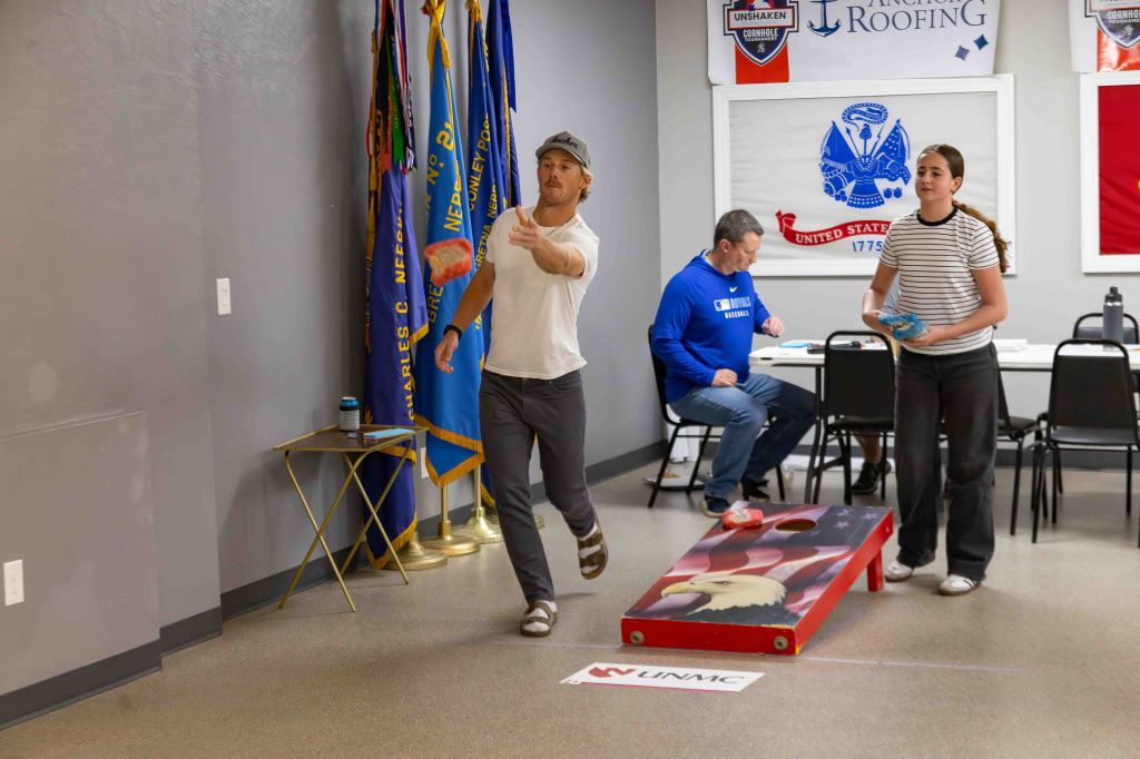 A group of people engaging in a cornhole game indoors, with an American flag-themed board and bags. A man in a white shirt is throwing a bag, while a woman in striping prepares to throw another. A third person is seated at a table in the background.