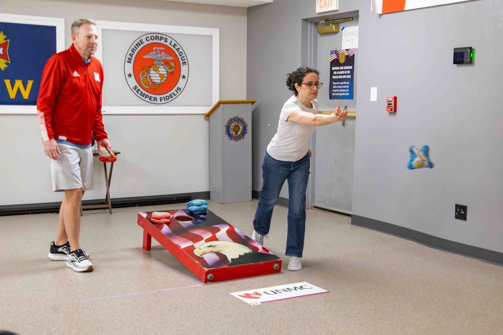 A woman playing bean bag toss, aimed at a slanted board, while a man stands nearby holding a bean bag, in a recreational room decorated with flags and a Marine Corps emblem.