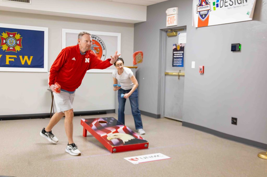 A man in a red shirt and shorts throws a bean bag towards a cornhole board, while a woman in a white shirt and jeans prepares to throw another bean bag in a recreational setting.