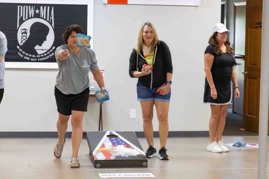 Participants playing a bean bag toss game in a recreational space, with one person throwing a bag, while others observe.