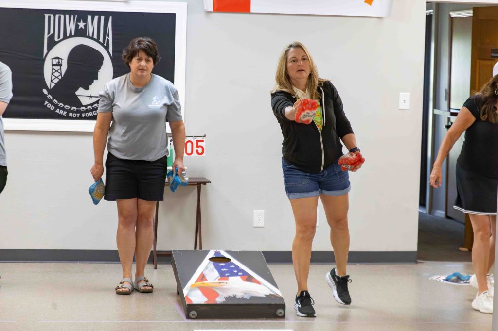 A group of women participating in a bean bag tossing game indoors, with one woman in the foreground throwing a bag while others observe. A POW/MIA banner is displayed on the wall behind them.