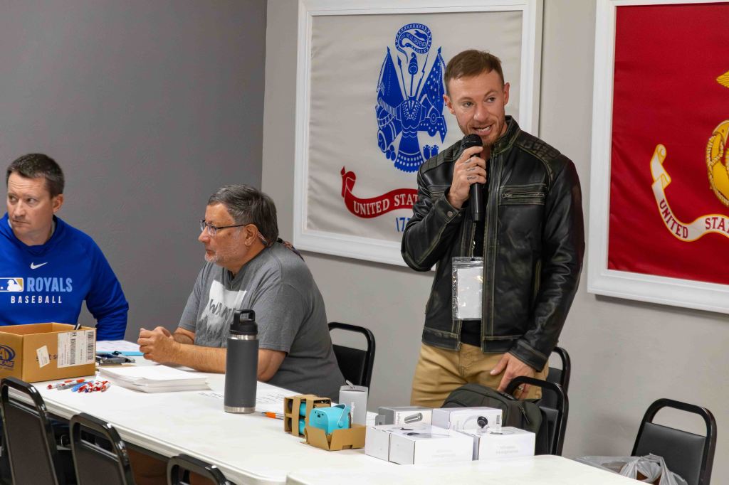 Three men at a table during an event, one is speaking into a microphone while the others listen. Items are placed in front of them, with flags displayed on the wall behind.