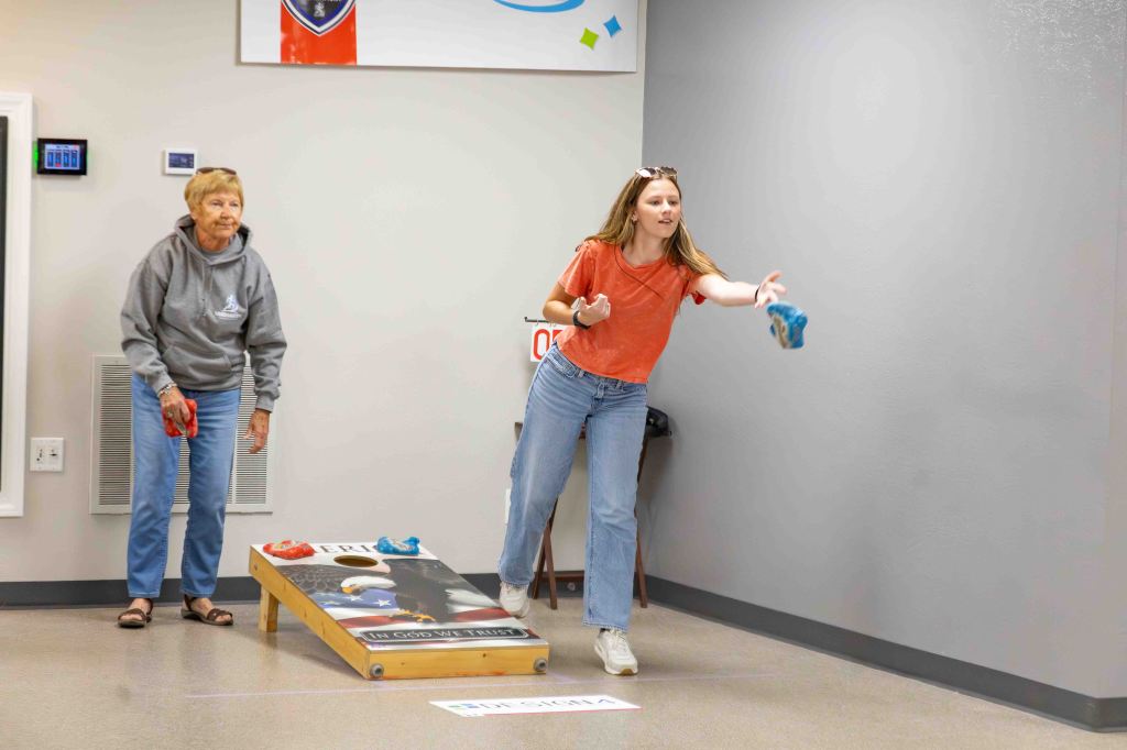A young woman wearing an orange shirt and blue jeans is throwing a blue bean bag towards a cornhole board. An older woman in a gray hoodie observes, holding a red bean bag, in an indoor setting.