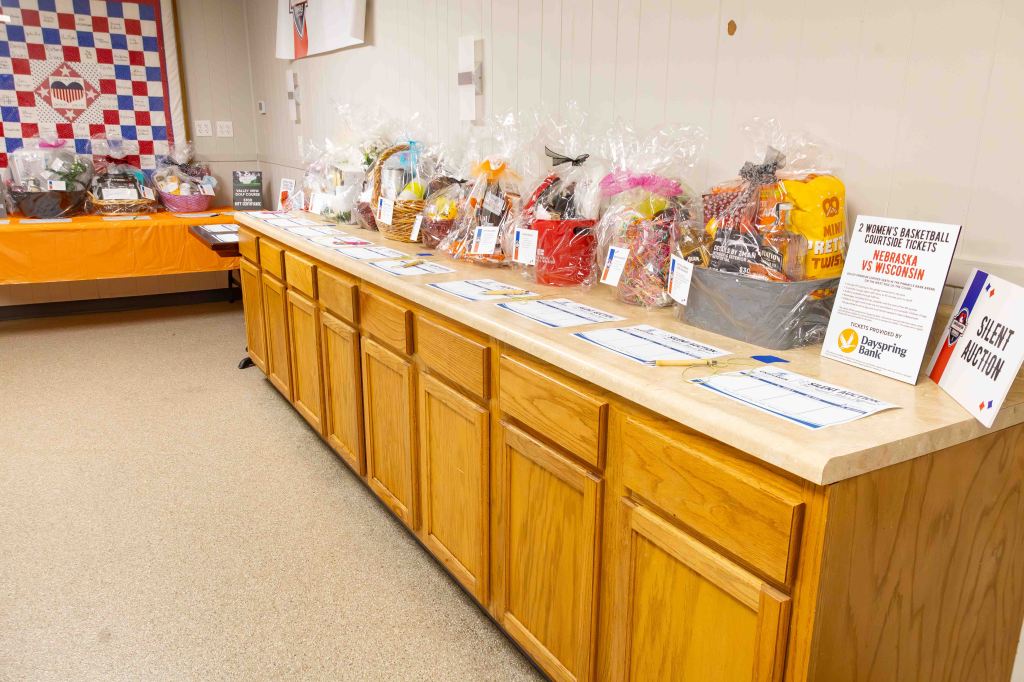 A wooden countertop displaying various gift baskets for a silent auction, with labeling cards for each item, set in a room with a quilted wall hanging in the background.