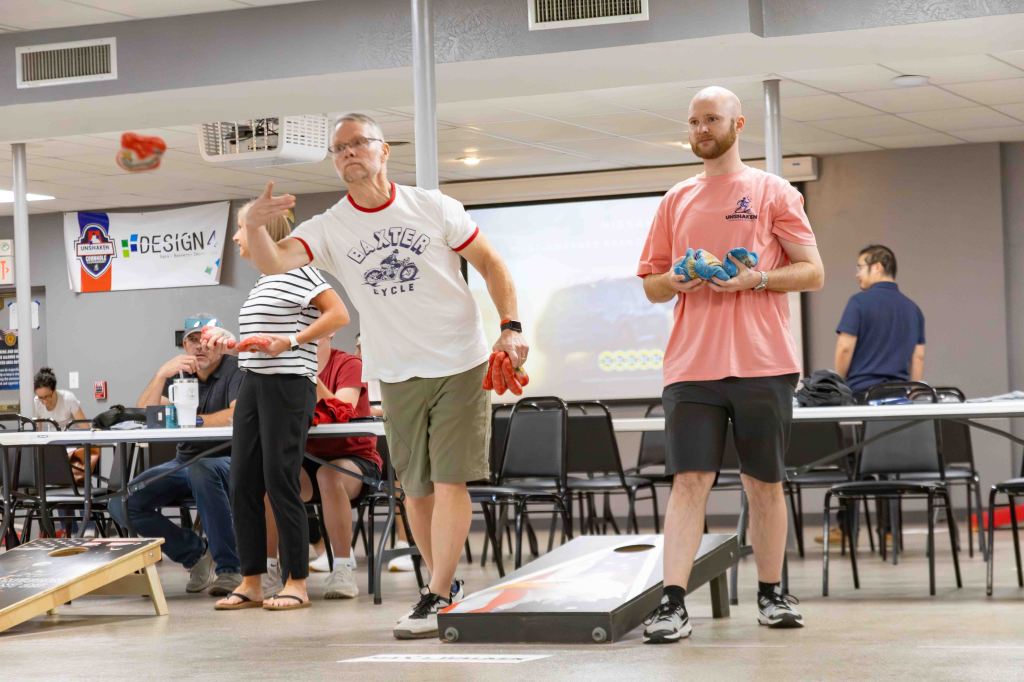 Two men playing a game of cornhole in a casual indoor setting, with tables and people in the background.