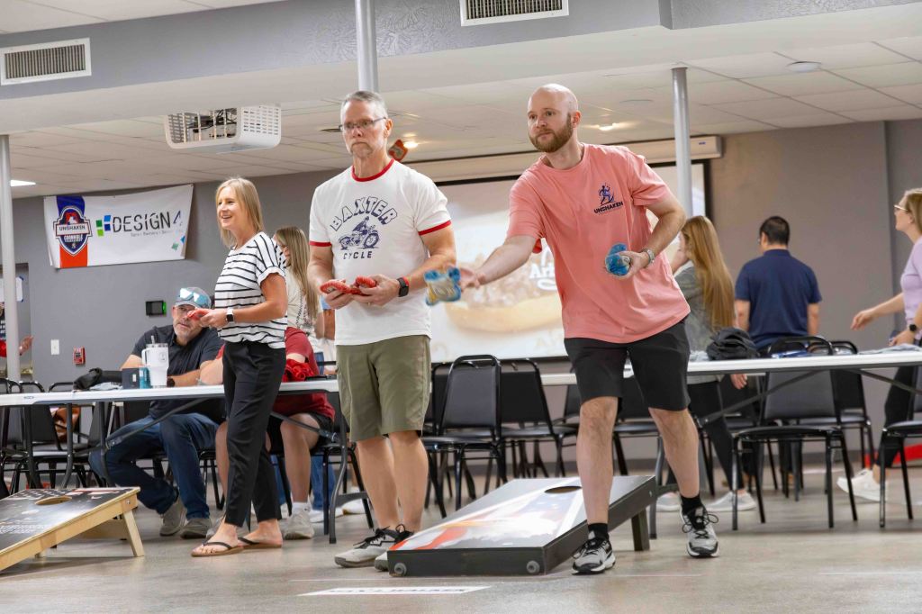 A group of people playing cornhole at an indoor event, with two players actively throwing bags towards a target board, while others watch from a table.