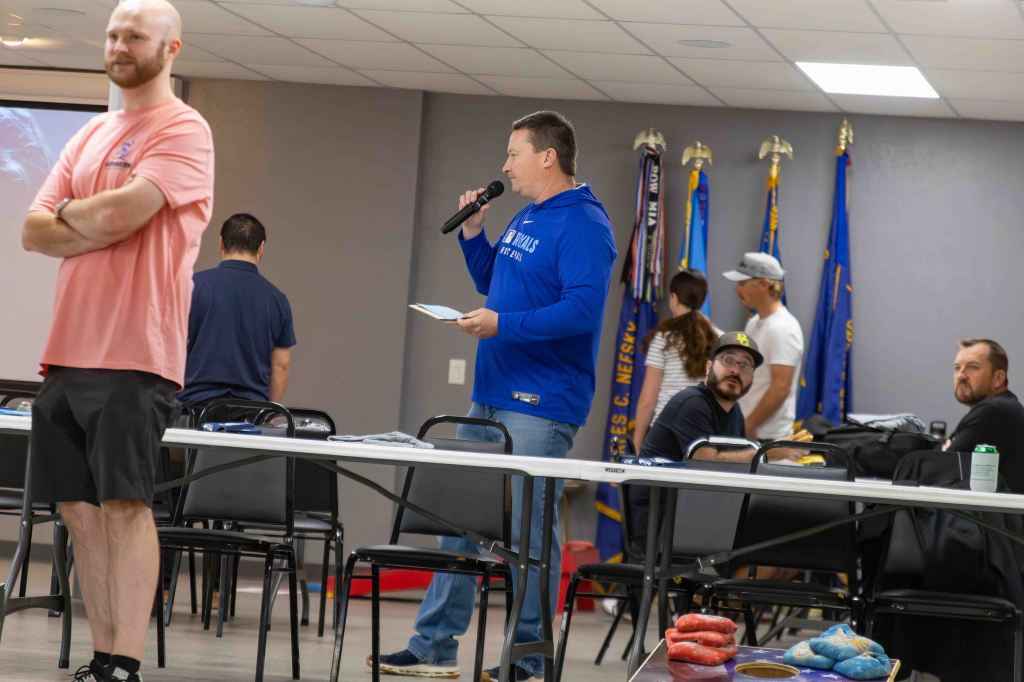 A man speaking into a microphone while holding notes in a meeting room, with several people listening and various flags displayed in the background.
