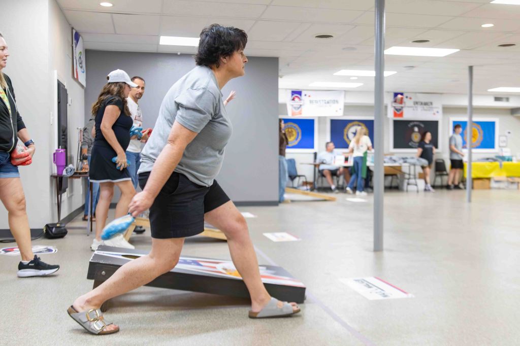 A group of people participating in a cornhole game at an indoor event, with one woman in the foreground throwing a bean bag while others watch and tables are set up in the background.