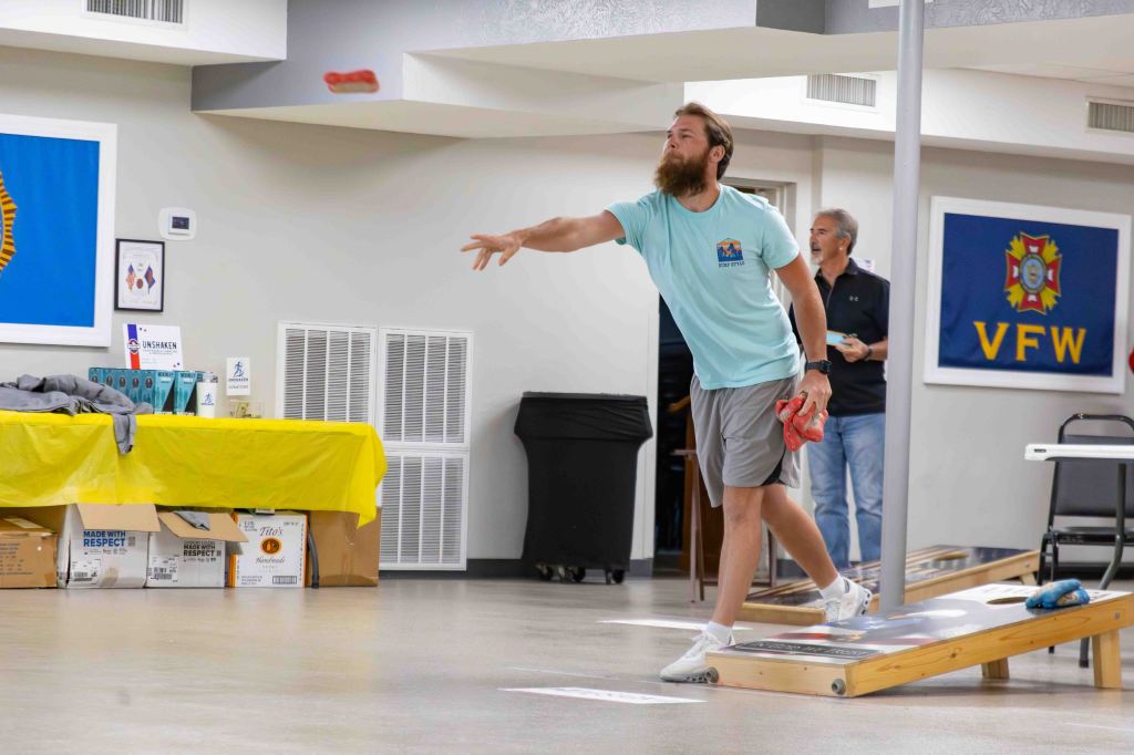 A man with a beard in a blue t-shirt is throwing a bean bag towards a cornhole board in an indoor setting, with a table in the foreground and other people in the background.
