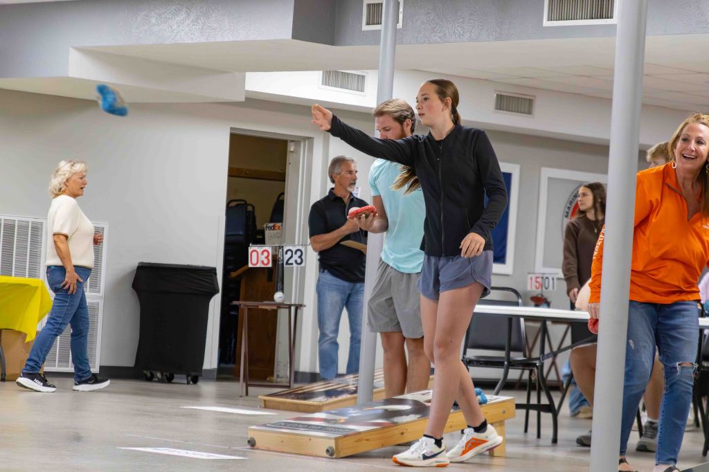 Group of people participating in a bean bag toss game indoors, some watching while others play.