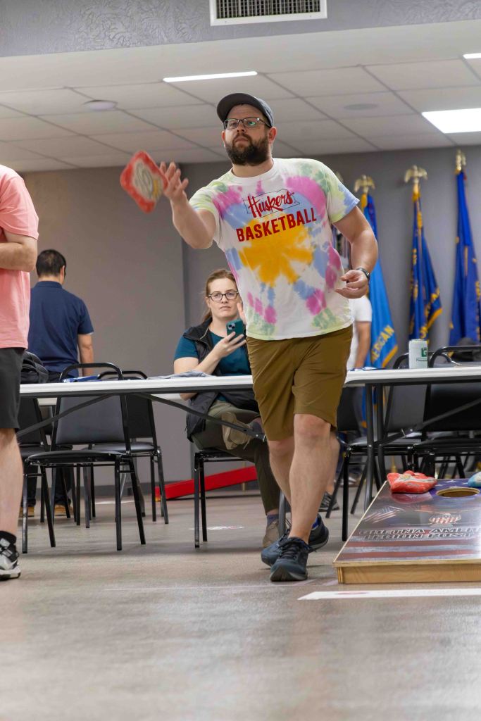 A man wearing a colorful tie-dye shirt and shorts throws a disc in a casual indoor setting with tables and chairs, while other people watch.
