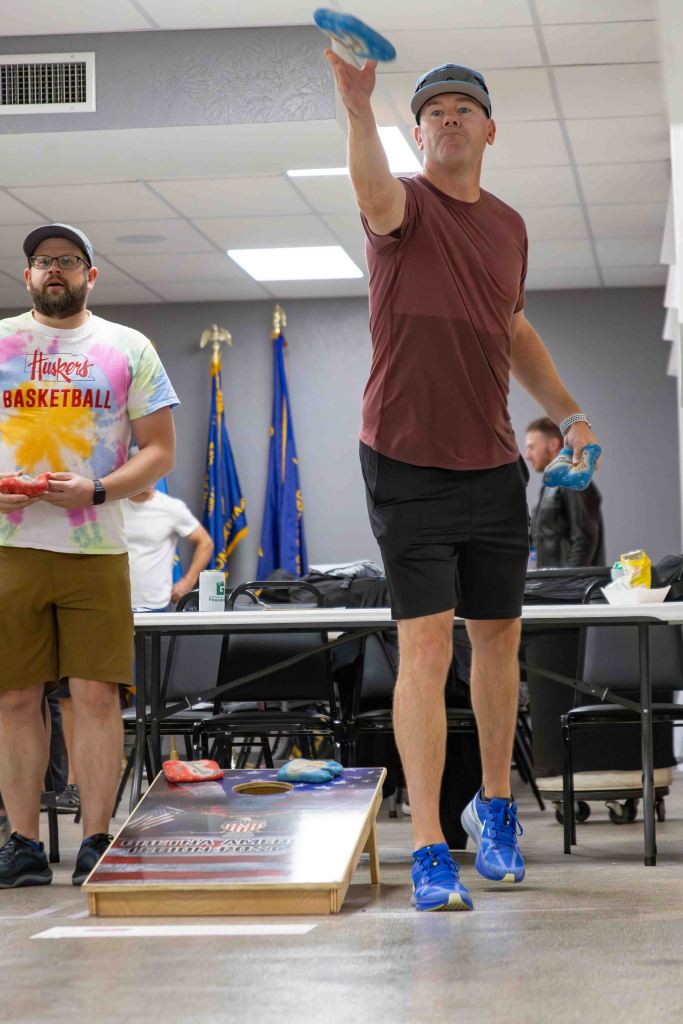 A man in a maroon shirt and black shorts is throwing a blue disc towards a cornhole board, while another man in a colorful tie-dye shirt stands nearby.