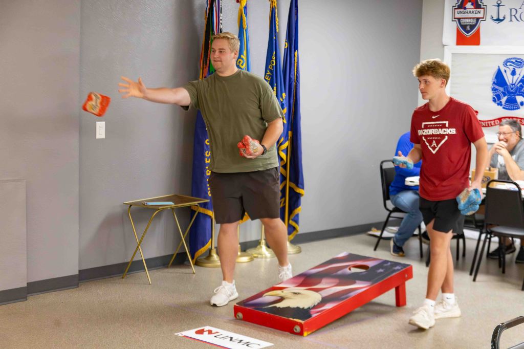 Two young men playing cornhole in a community center, one throwing a bean bag while the other waits to throw.