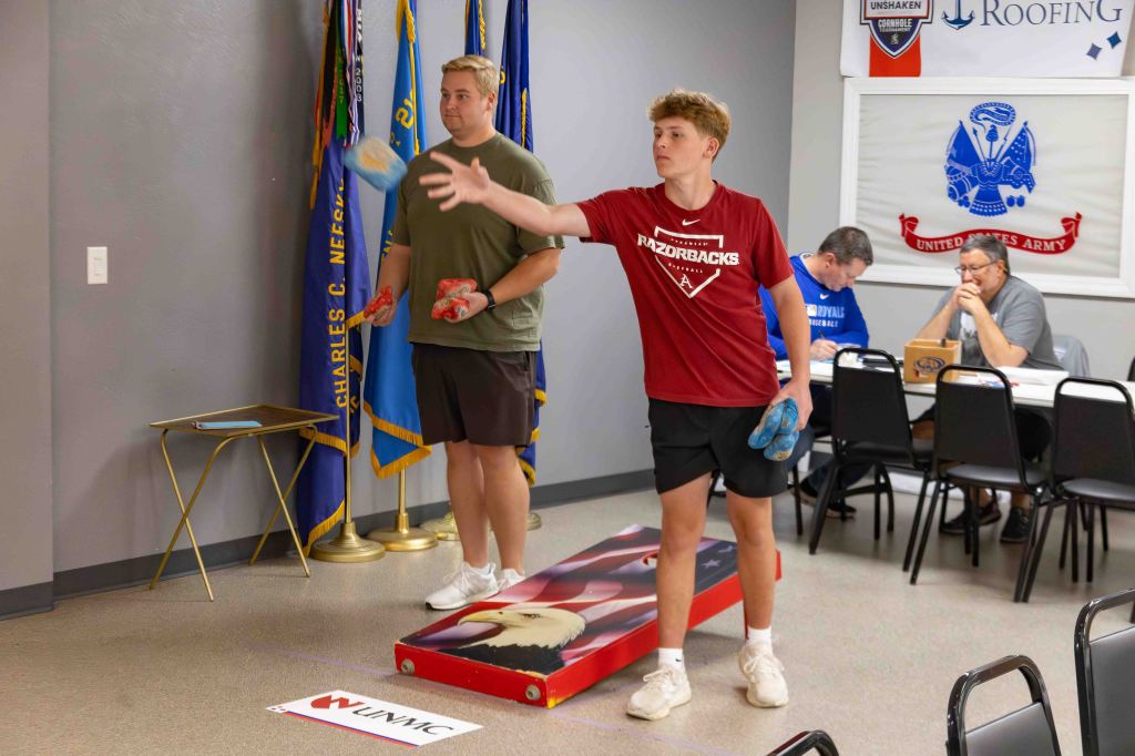 Two young men participating in a cornhole game indoors, one about to throw a bag while the other watches. In the background, people are seated at a table.