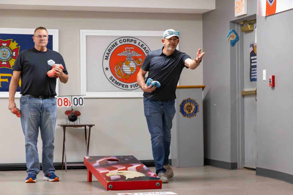 Two men playing cornhole in a recreational space, with one man tossing a blue bean bag while the other stands holding a red bean bag. A scoreboard shows the score.