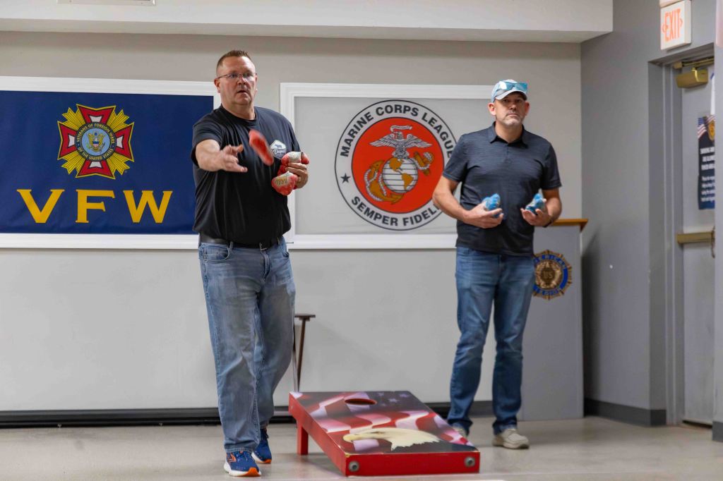 Two men participating in a bean bag tossing game indoors, with a VFW banner and Marine Corps League emblem in the background.