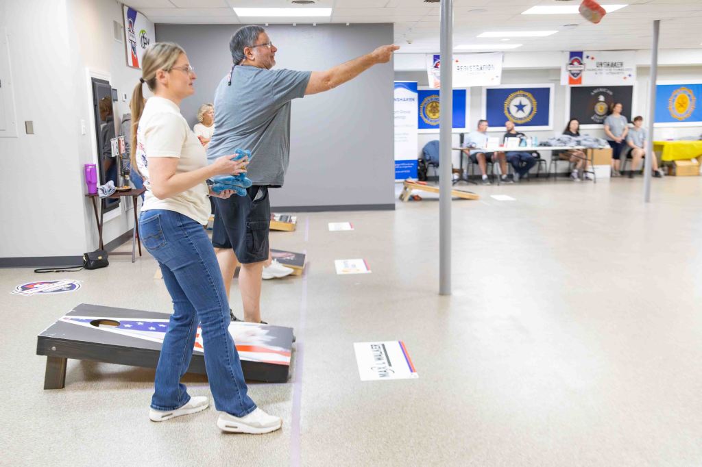 Two people participating in a cornhole game indoors, with several bean bags in the air and others waiting to be thrown. Several spectators are seated at tables in the background.