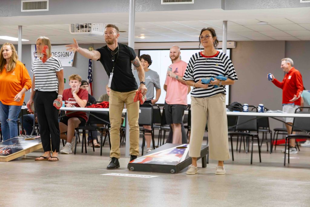A group of people participating in a cornhole game indoors, with players throwing bags towards the boards. Participants include both men and women, and there's a mix of expressions showing concentration and enjoyment.